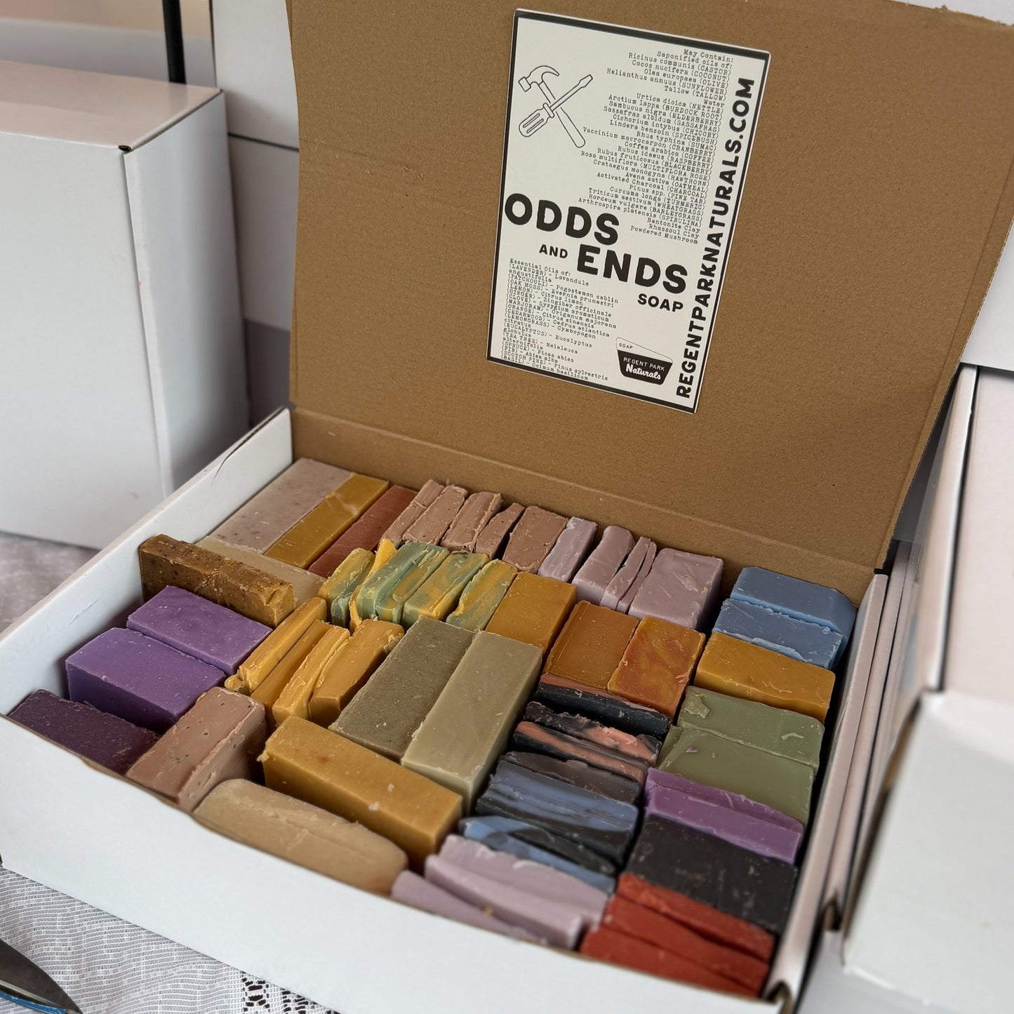 An open white box filled with many rectangular bars of soap in assorted earthy and pastel colors. The inside lid has a printed label reading 'ODDS AND ENDS SOAP' and 'REGENTNATURALS.