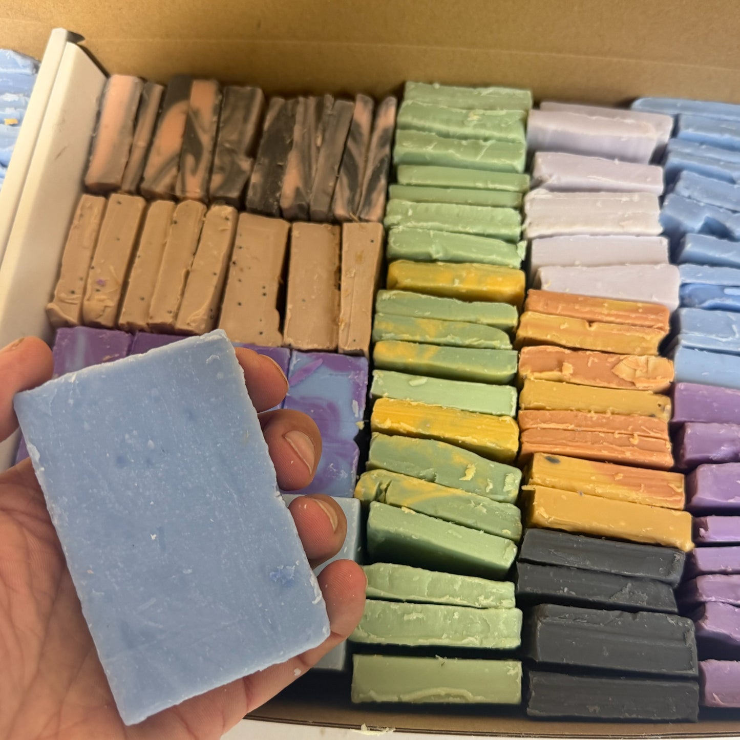 A hand holds a light blue rectangular bar of soap in front of an open cardboard box filled with many other colorful soap bars arranged in rows.
