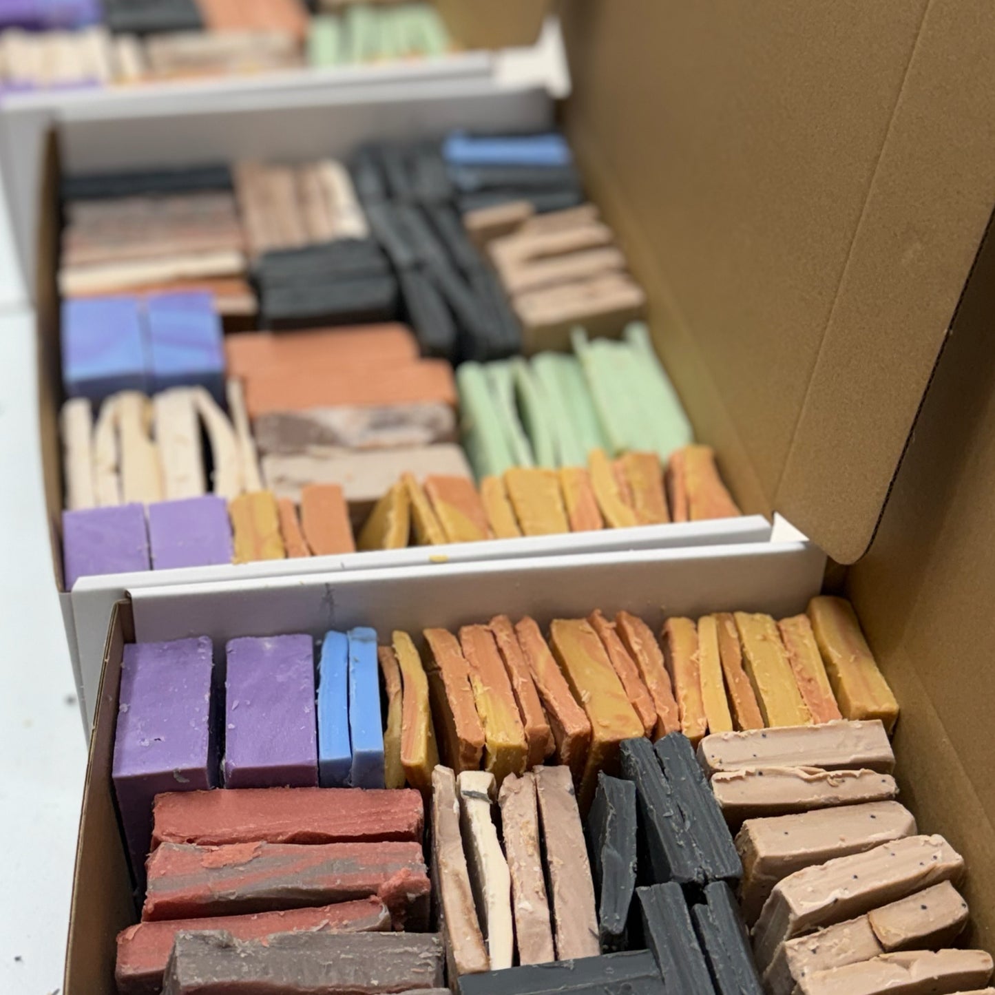 Open cardboard boxes filled with many small rectangular soap blocks of different colors, including purple, blue, orange, yellow, green, black, and beige, arranged in rows.