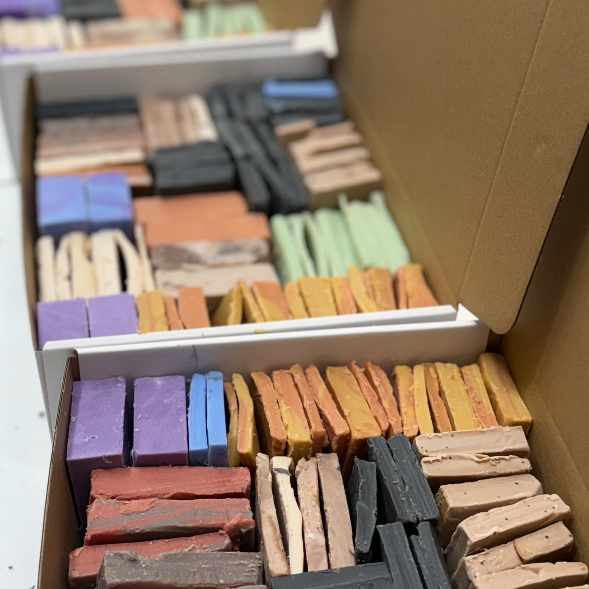 Open cardboard boxes filled with many small rectangular soap blocks of different colors, including purple, blue, orange, yellow, green, black, and beige, arranged in rows.