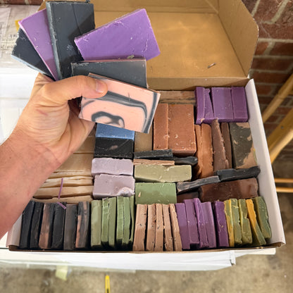A hand holds a pink and black swirled soap bar in front of an open cardboard box filled with many other colorful handmade soap bars in purple, green, brown, gray, and cream.