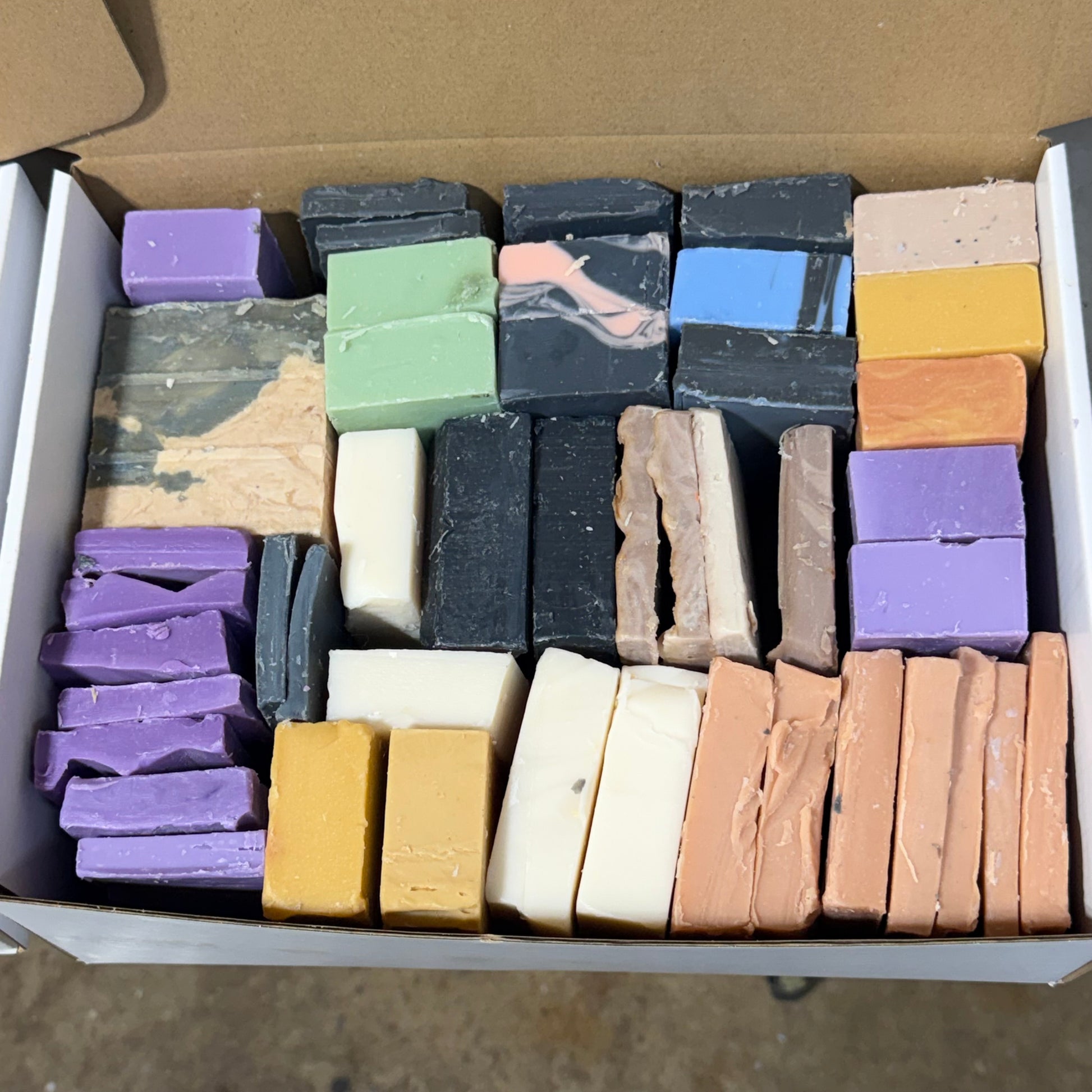 An open cardboard box filled with multiple rectangular bars of soap in assorted colors, including purple, green, black, blue, yellow, beige, brown, and cream, arranged in rows.